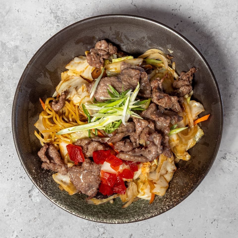 Stir-fried noodles with sliced beef, cabbage, peppers, carrots, and scallions served in a dark bowl.