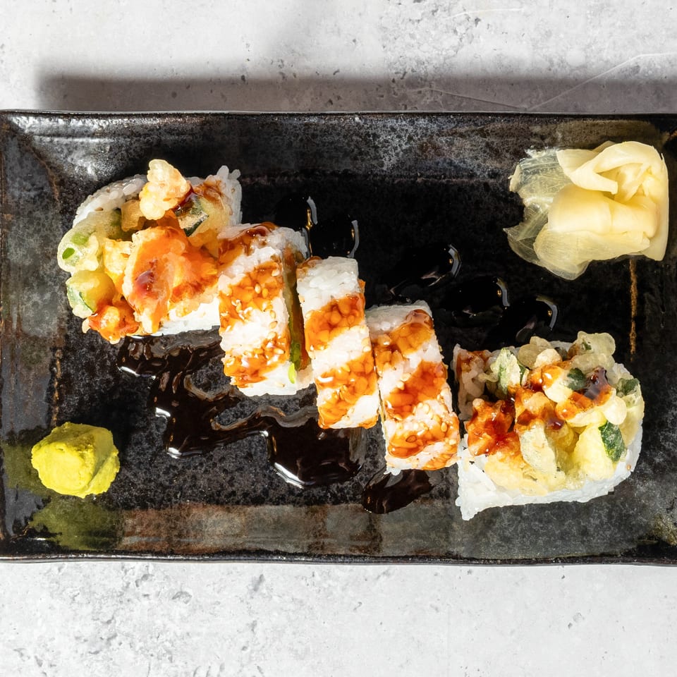 Assorted tempura sushi rolls drizzled with sauce on a black rectangular plate, served with pickled ginger and wasabi.