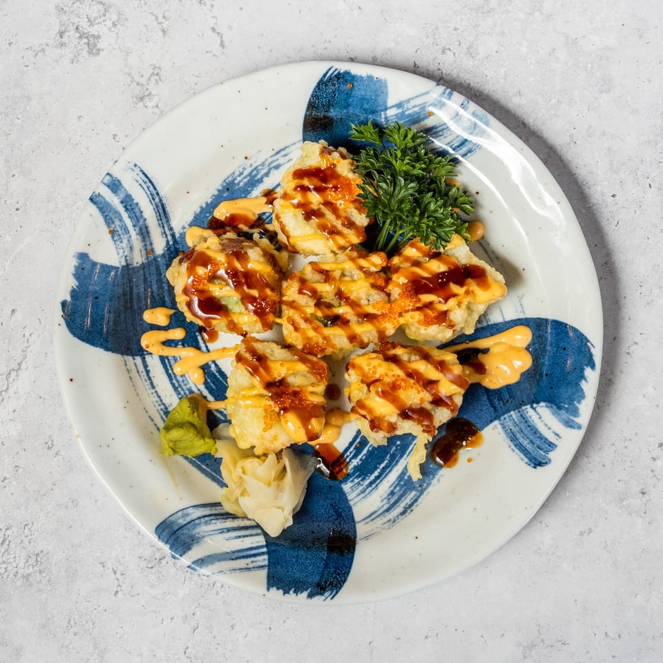 Crispy sushi rolls drizzled with spicy mayonnaise and eel sauce, garnished with roe and parsley on a blue-patterned plate.