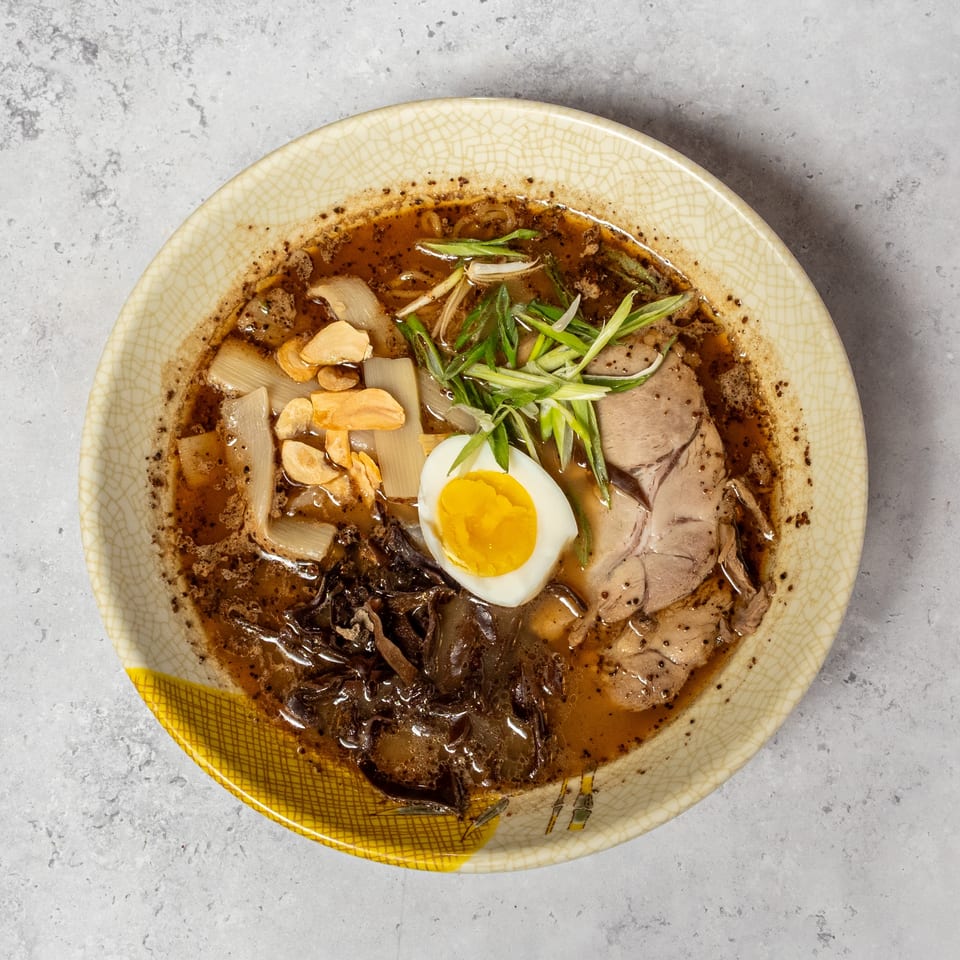 Bowl of dark broth ramen topped with sliced pork, soft-boiled egg, scallions, garlic, bamboo shoots, and mushrooms.