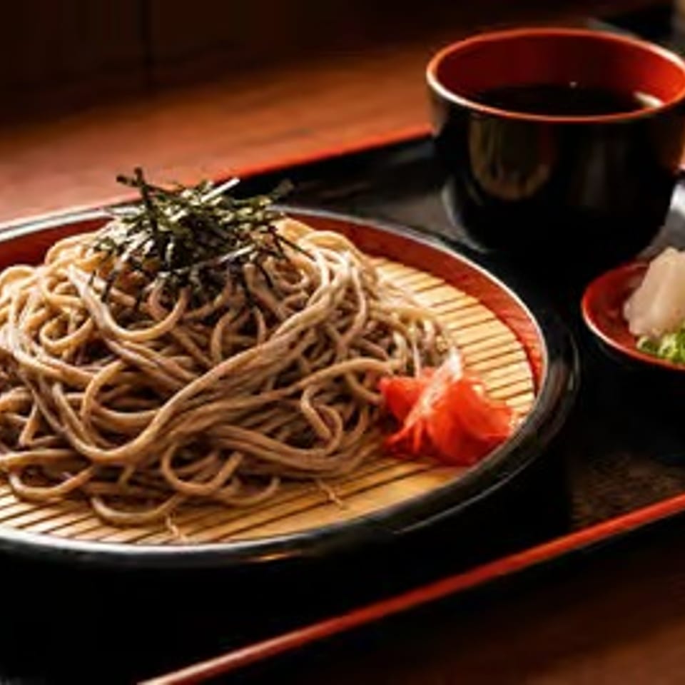 Japanese soba noodles topped with shredded seaweed, served with dipping sauce and garnishes on a black tray.