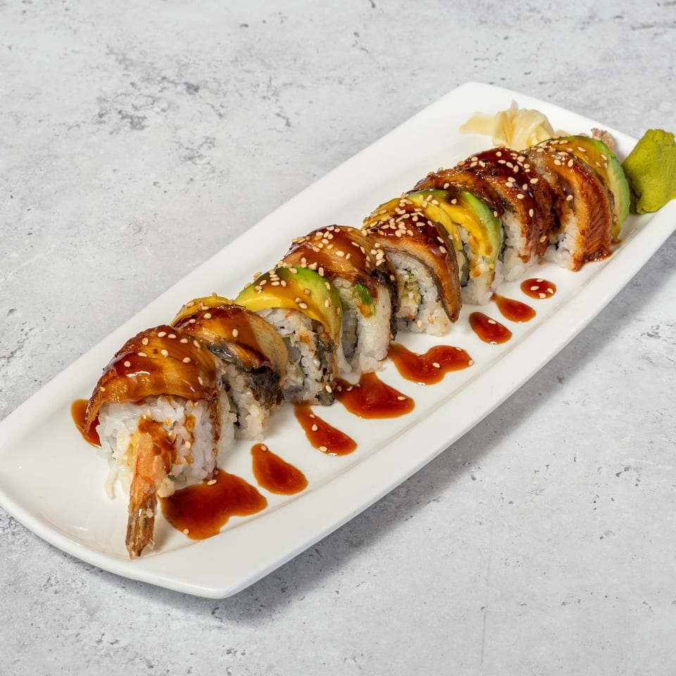 Sushi roll slices topped with glazed eel and sesame seeds, accompanied by avocado, pickled ginger, wasabi, and sauce.