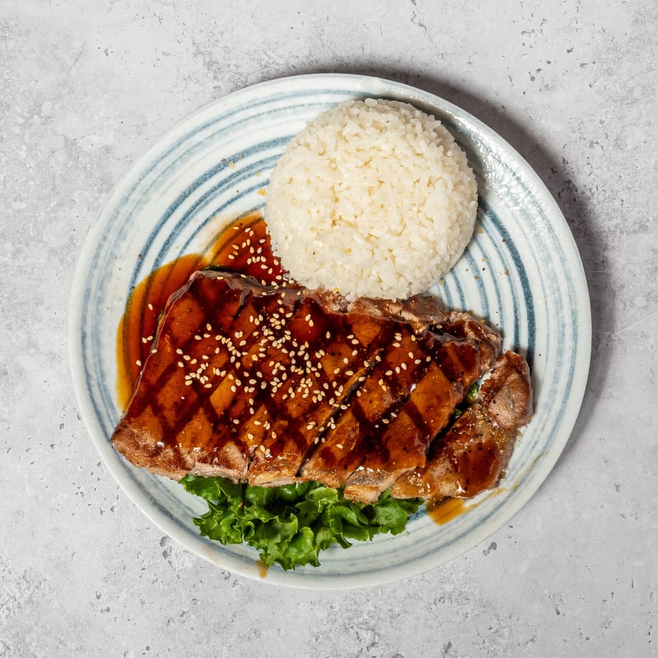 Glazed sliced meat topped with sesame seeds, served with white rice and leafy greens on a striped plate.